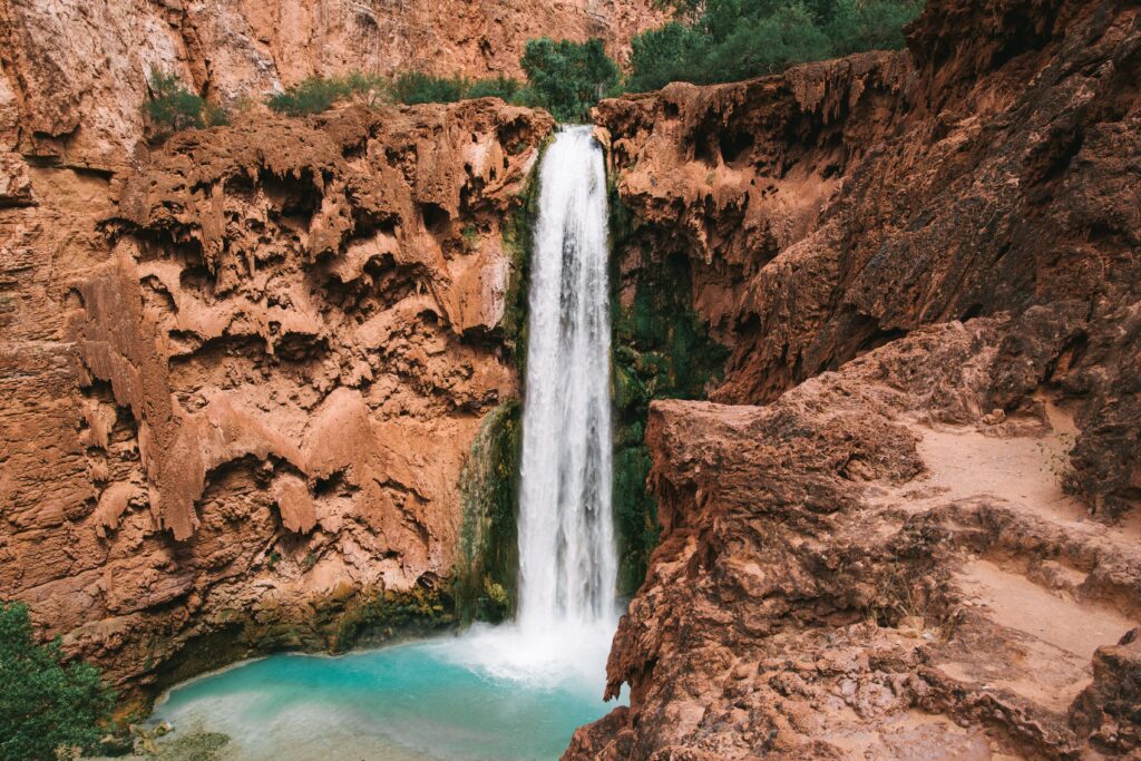 Waterfall cascading over red rocks into turquoise pool surrounded by lush greenery and rocks Wallpaper