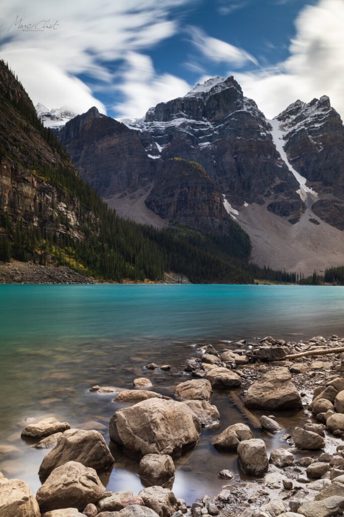 Turquoise river flowing past smooth stones with mountains and evergreen forest under clouds Wallpaper