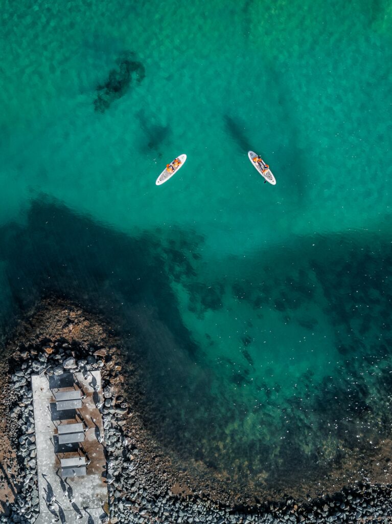 Aerial view of turquoise sea with two boats near a rocky coastline showing clear water and rocks Wallpaper