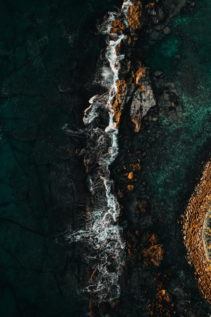 Aerial view of turquoise waves crashing on sunlit rugged rocks nature wallpaper background