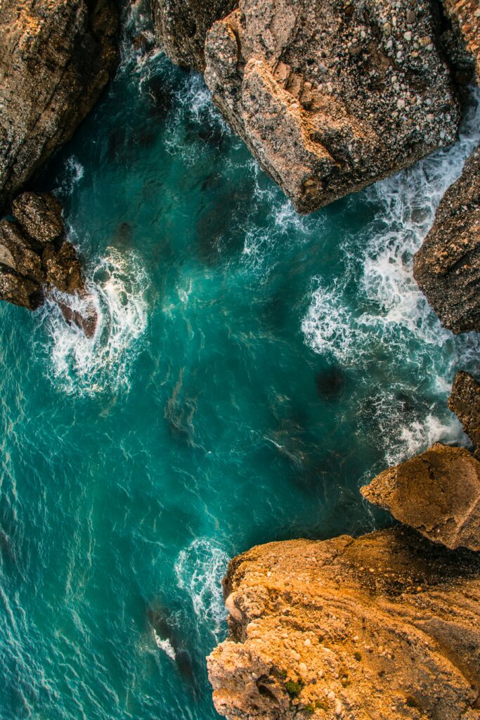 Aerial view of rugged cliffs framing vibrant turquoise sea with foamy waves crashing coast Wallpaper