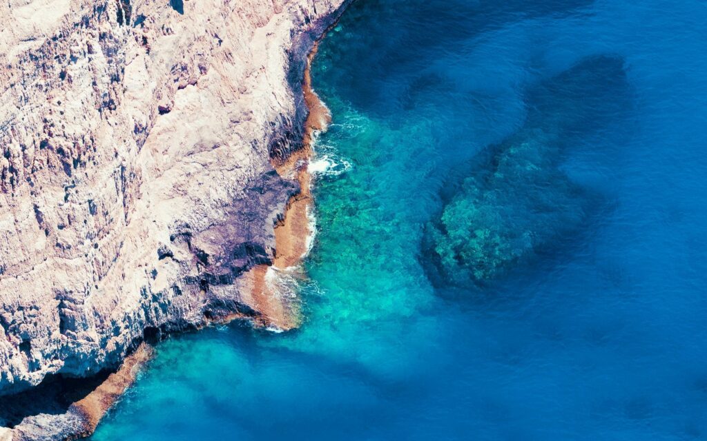 Aerial view of turquoise sea meeting rugged beige rocky coastline with sunlit clear shallow beach Wallpaper