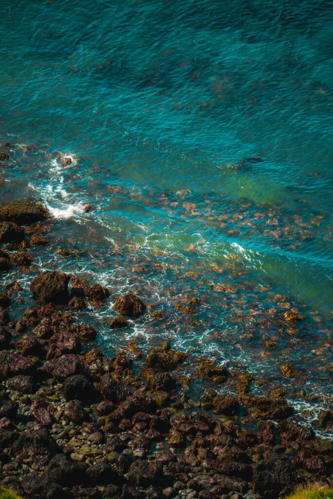 Turquoise sea waves crashing over dark rugged stones with foamy surf in natural sunlight wallpaper
