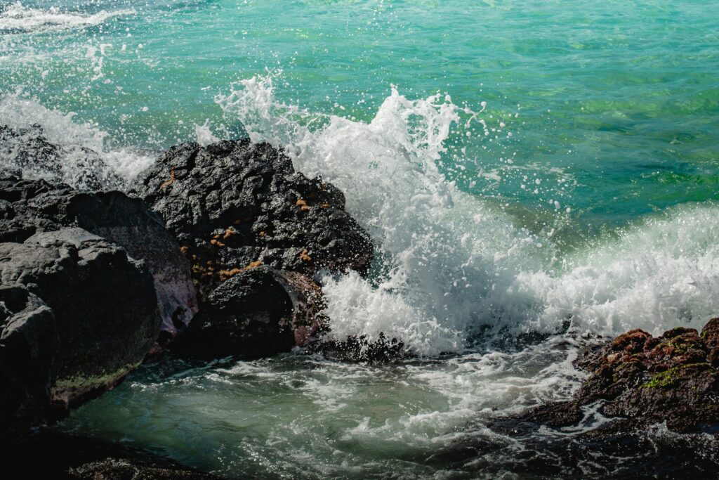 Turquoise sea waves crash on dark rugged rocks with white surf sprays coastal landscape view Wallpaper