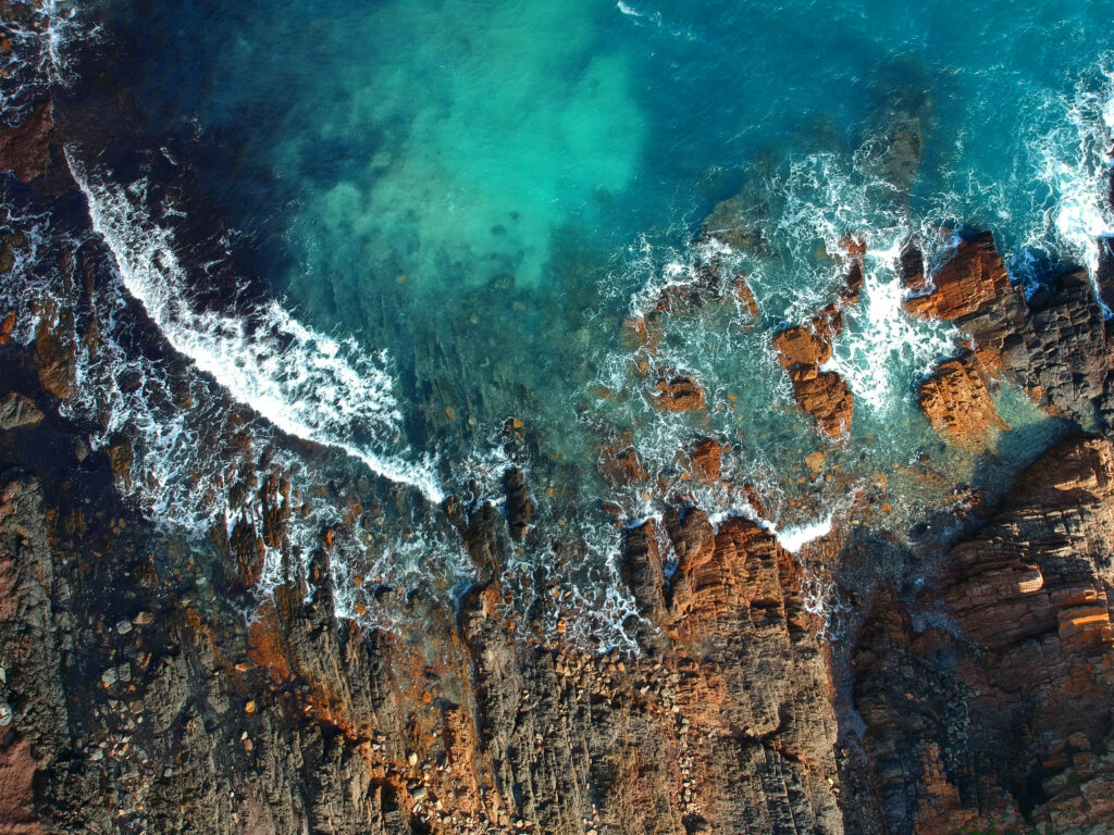 Aerial view of rugged rocks reefs and turquoise sea with foamy waves along the coastline Wallpaper