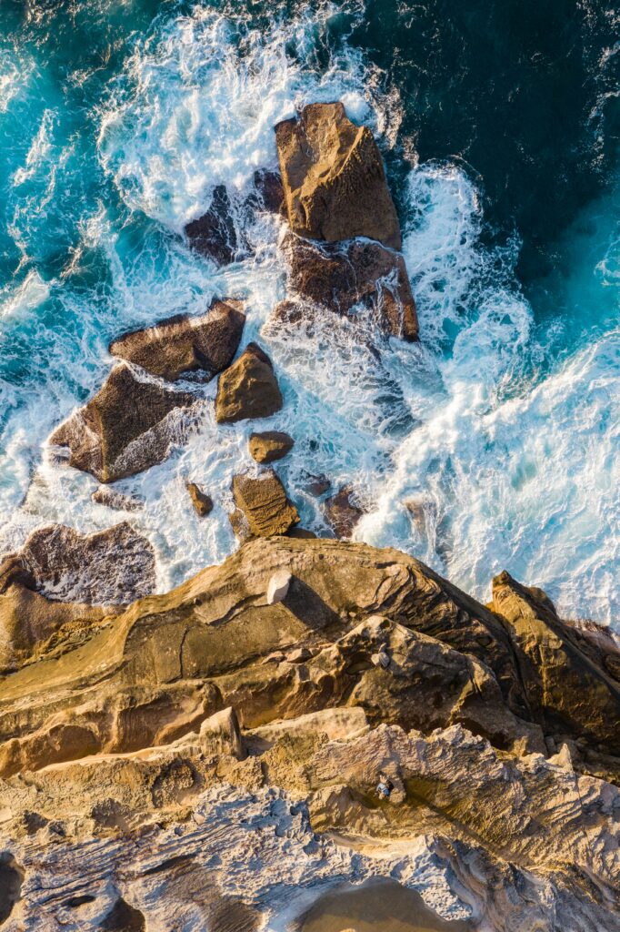 Aerial view of rugged rocky shoreline with turquoise waves crashing creating dynamic surf energy Wallpaper