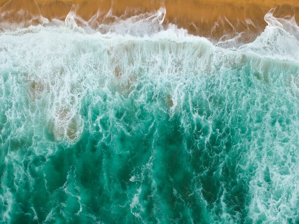 Aerial view of turquoise sea waves crashing on sandy beach with vibrant foam and sand textures Wallpaper