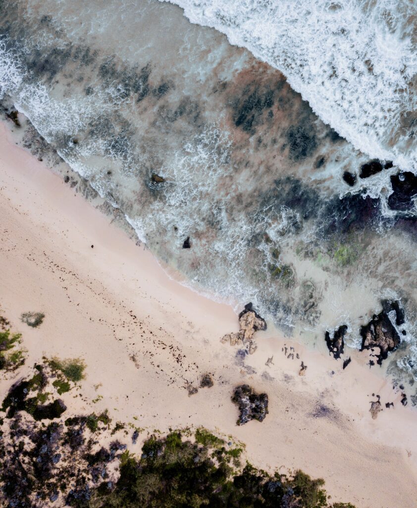 Aerial view of turquoise waves meeting beige sand on rugged beach with rocks and green plants Wallpaper