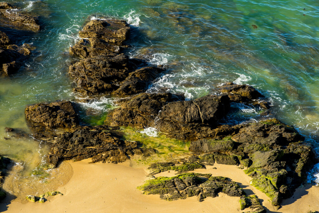 Turquoise sea waves crashing dark rugged reefs on golden sandy shore coastal wallpaper background