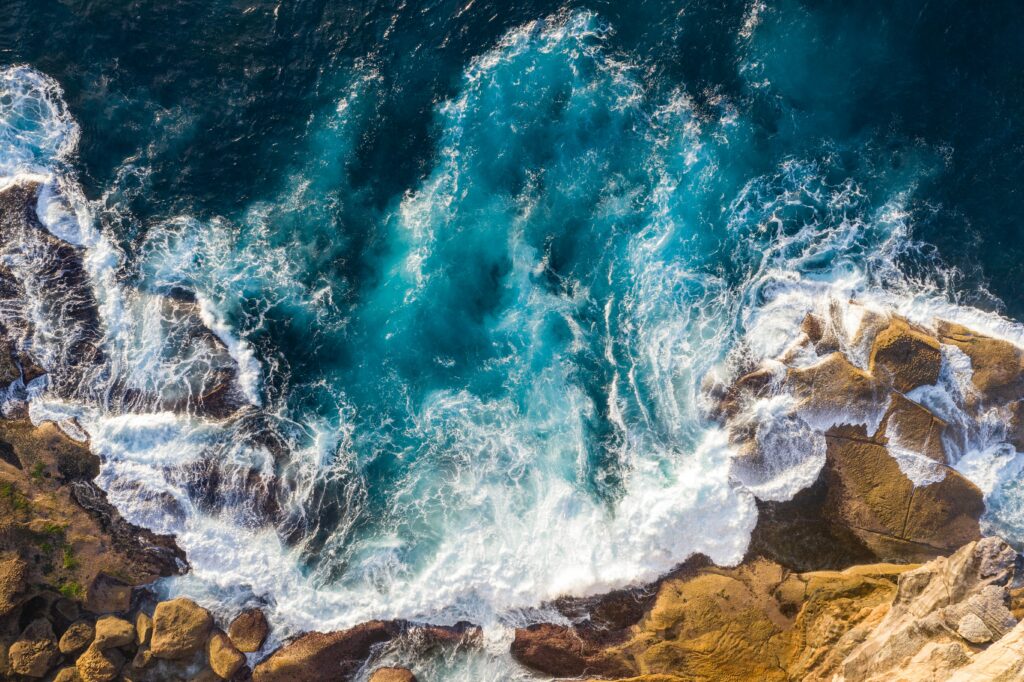 Aerial view of turquoise sea crashing on rugged rocky shore with foamy swirling waves wallpaper