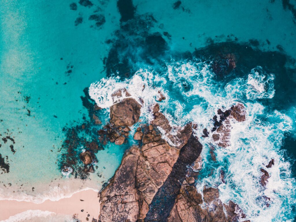 Aerial view of turquoise sea waves crashing against rugged rocky shore creating white foam patterns Wallpaper