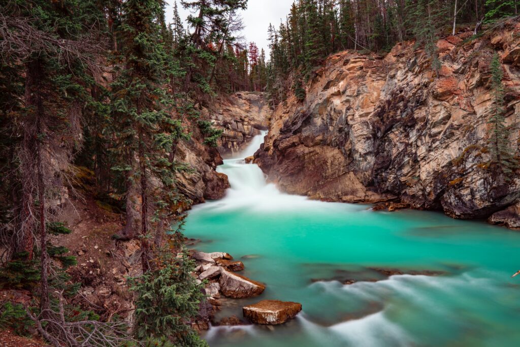 Turquoise waterfall flowing over rocks between rugged cliffs and lush pine forest landscape wallpaper
