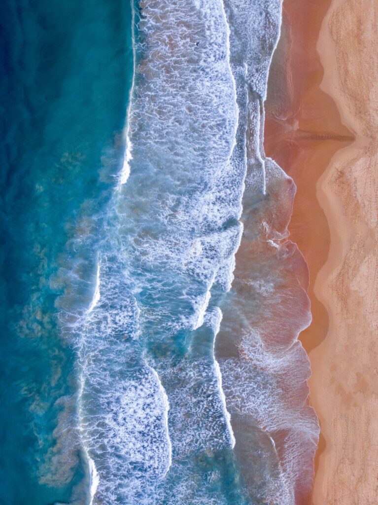 Aerial view of turquoise waves rolling onto sandy beach with deep blue water and white foam Wallpaper