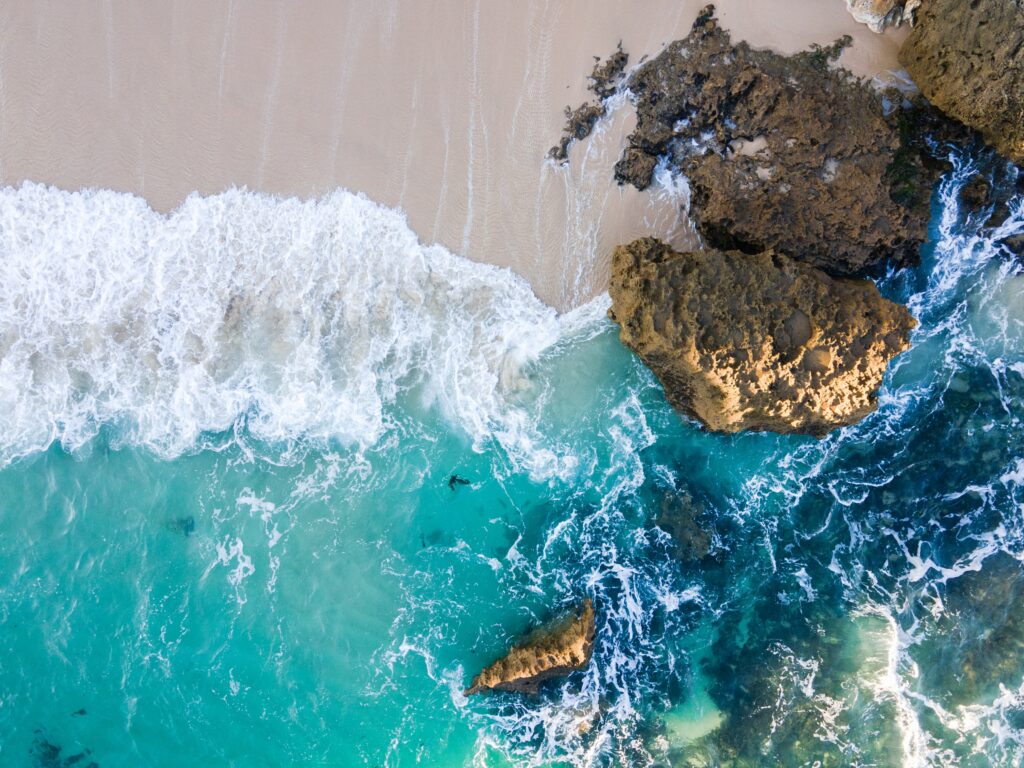 Aerial view of turquoise waves meeting sandy beach with rugged rocks coastal wallpaper background