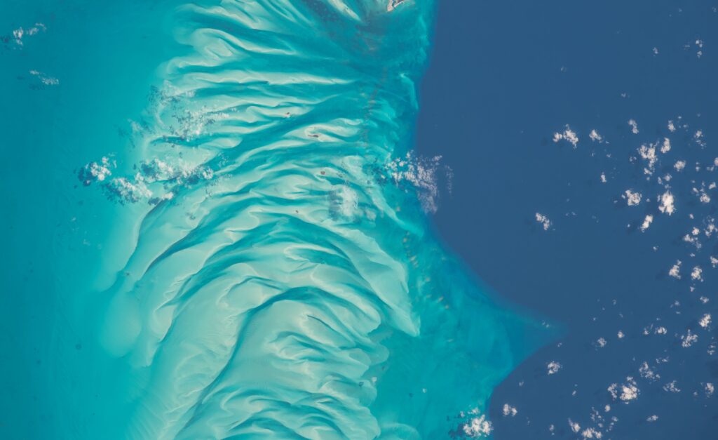 Aerial view of turquoise and deep blue sea with sandy patterns and scattered clouds wallpaper