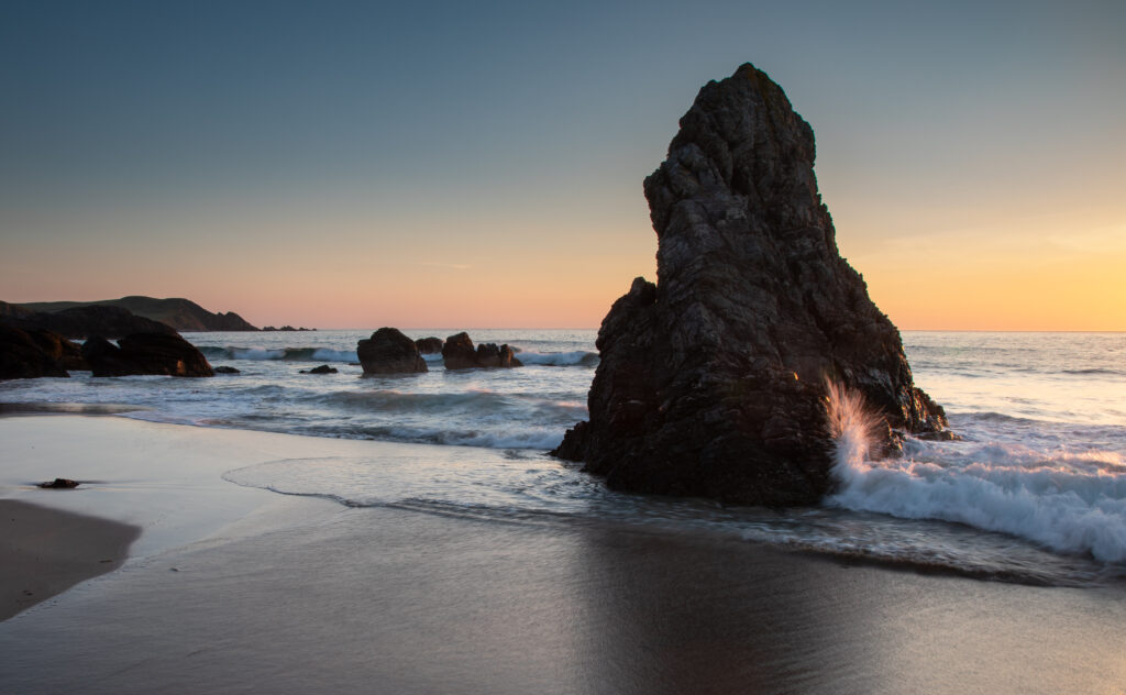 Twilight coastal landscape with rugged rocks gentle sea waves and warm golden blue horizon background Wallpaper