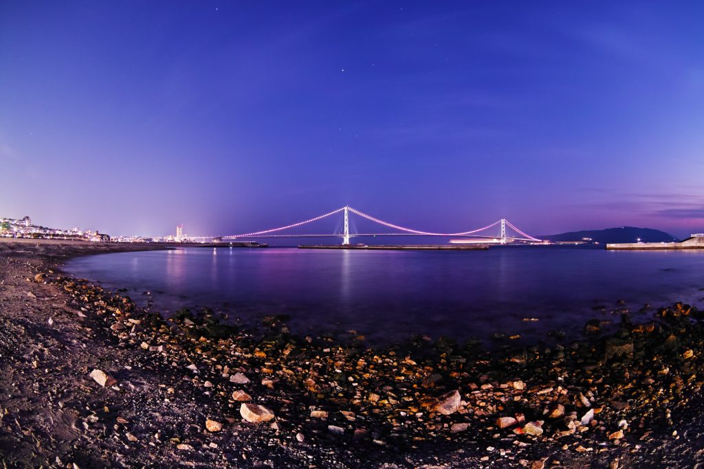 Twilight coastal wallpaper with smooth pebbles shells illuminated bridge and calm waters background