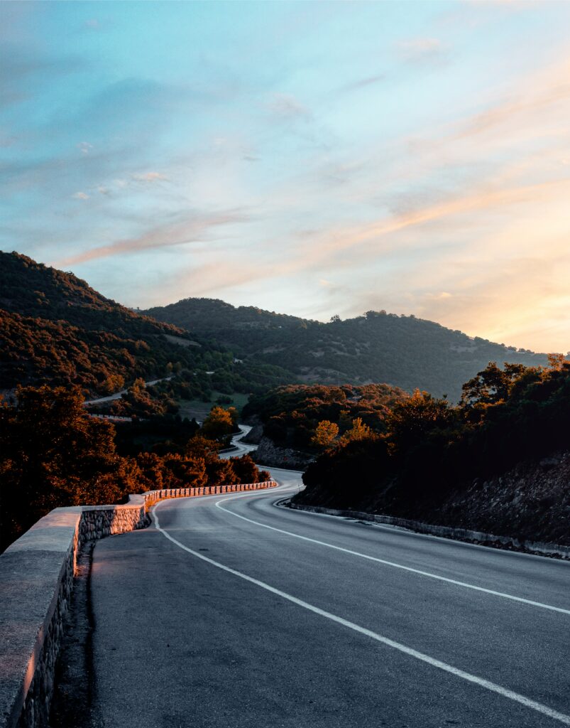 Winding mountain road through lush green trees under twilight sky with orange and blue hues Wallpaper