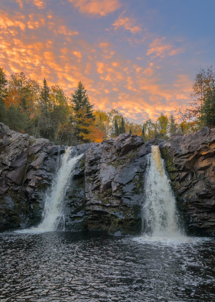 Twin waterfalls over rugged cliff into river surrounded by autumn trees under vibrant sunset sky Wallpaper
