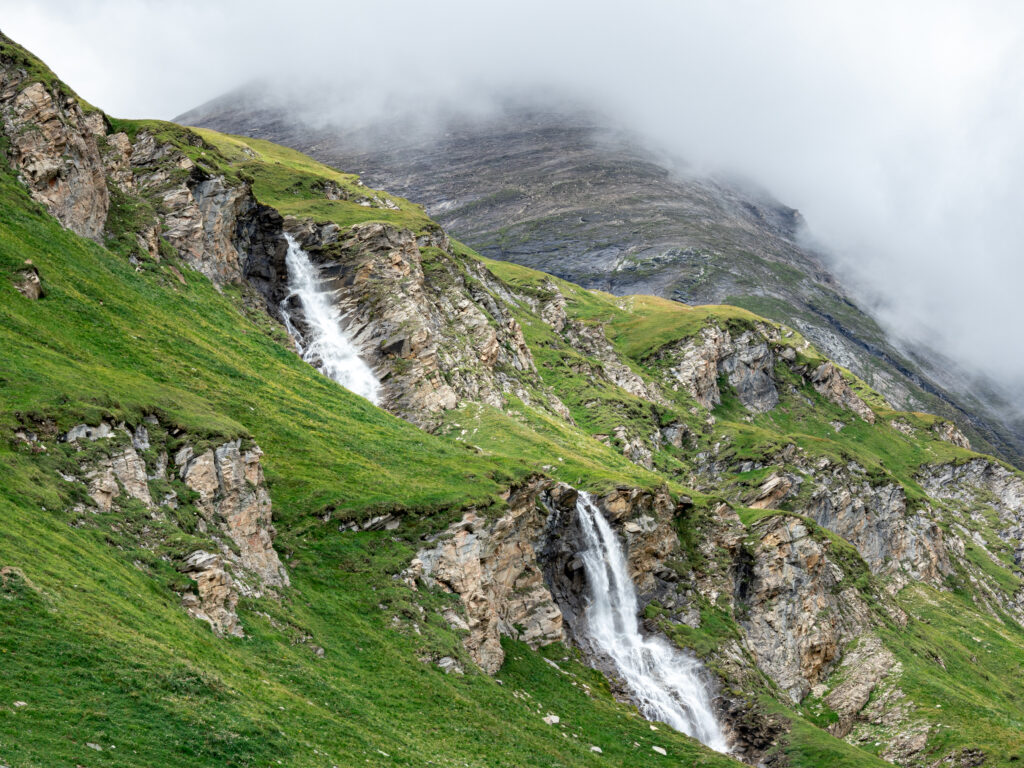 Twin waterfalls cascading rocky cliffs surrounded by green grass and misty mountain background Wallpaper