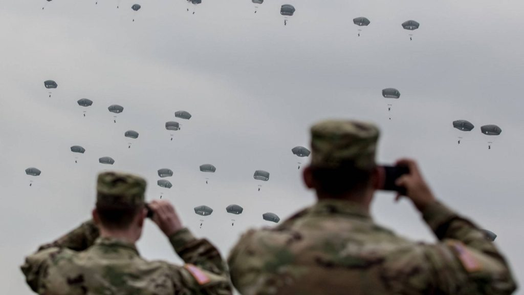 Two US Army soldiers in OCP uniforms watch massive airborne paratroopers descending under cloudy sky Wallpaper
