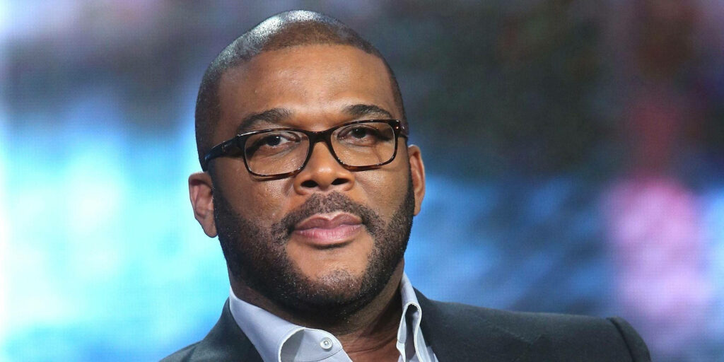 Tyler Perry close up with confident expression and signature glasses on blurred wallpaper background