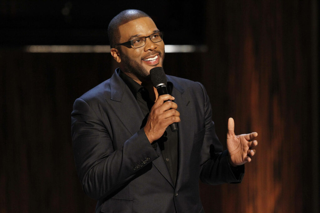Tyler Perry speaking on stage in black suit holding microphone with warm engaging expression HD Wallpaper