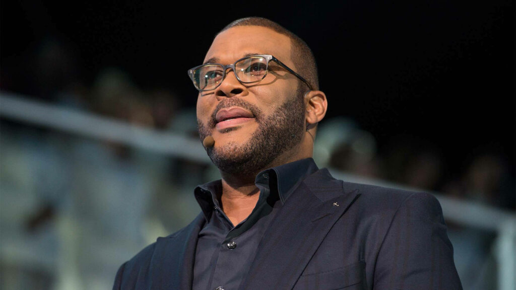 Tyler Perry delivering keynote speech in dark suit and glasses with dynamic focused background Wallpaper