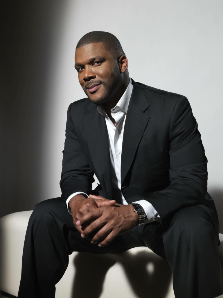 Tyler Perry in sleek black suit smiling confidently against a light minimalist wallpaper background