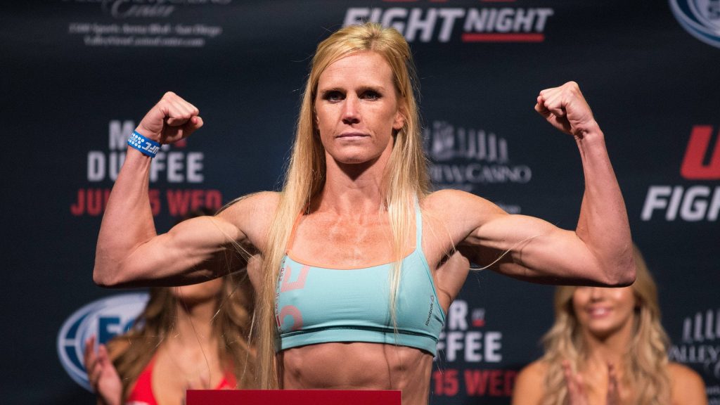 UFC champion Holly Holm flexing in blue sports top at dark Fight Night weigh-in background wallpaper