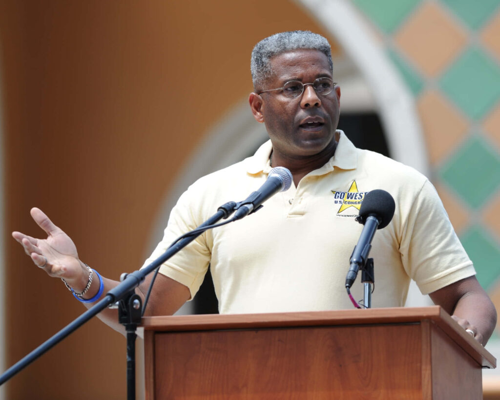 US Congressman Allen West speaking passionately at outdoor event podium colorful architecture background Wallpaper