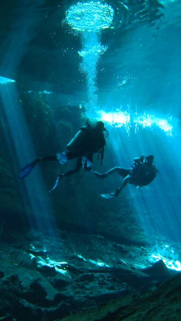 Two divers exploring a shimmering blue underwater cave with natural light rays and textures Wallpaper