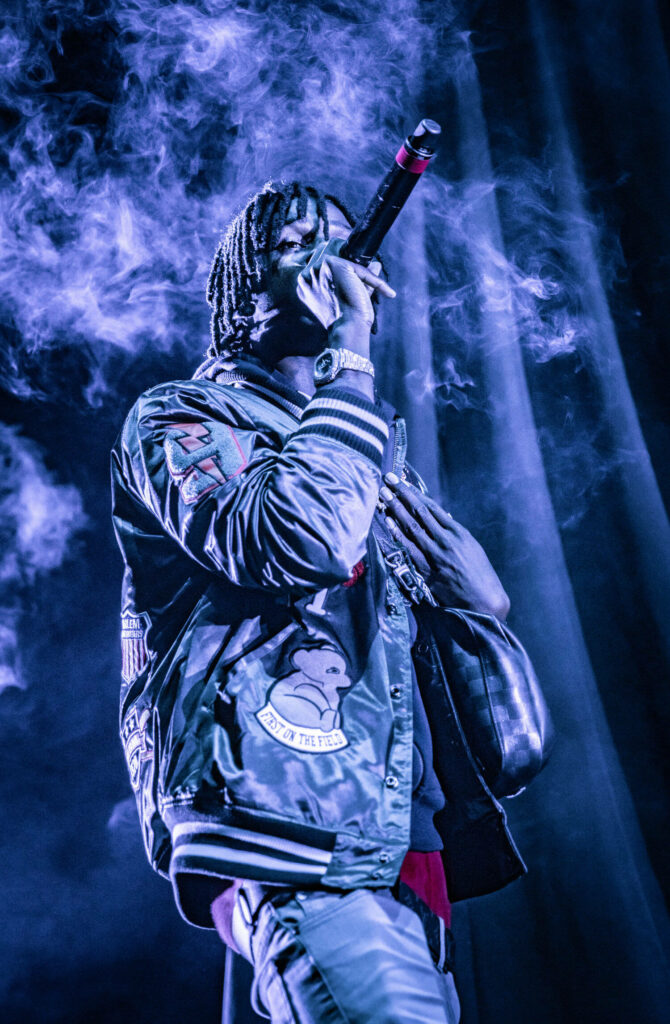 Chief Keef performing live onstage in purple lights wearing patch varsity jacket haze background Wallpaper