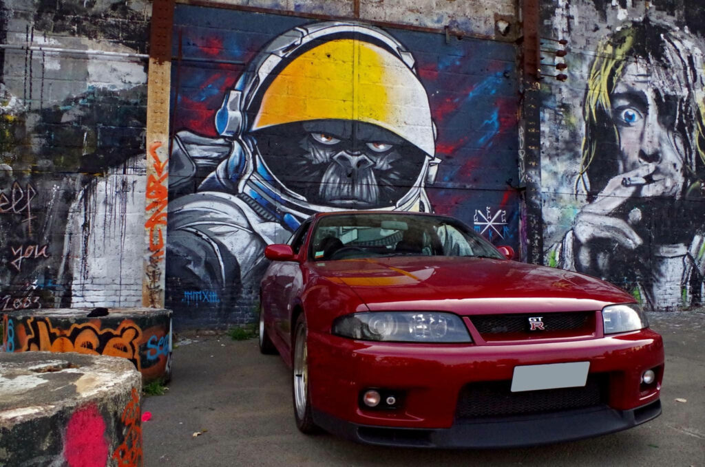 Cruising in Urban Cool: Nissan Skyline Gtr R33 Poses Confidently Against Vibrant Graffiti Street Art Wallpaper