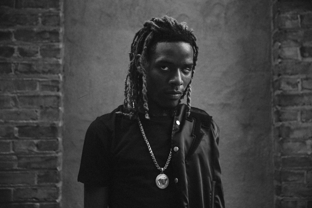 Rapper with dreadlocks and bold Versace Medusa necklace against textured brick wall background Wallpaper