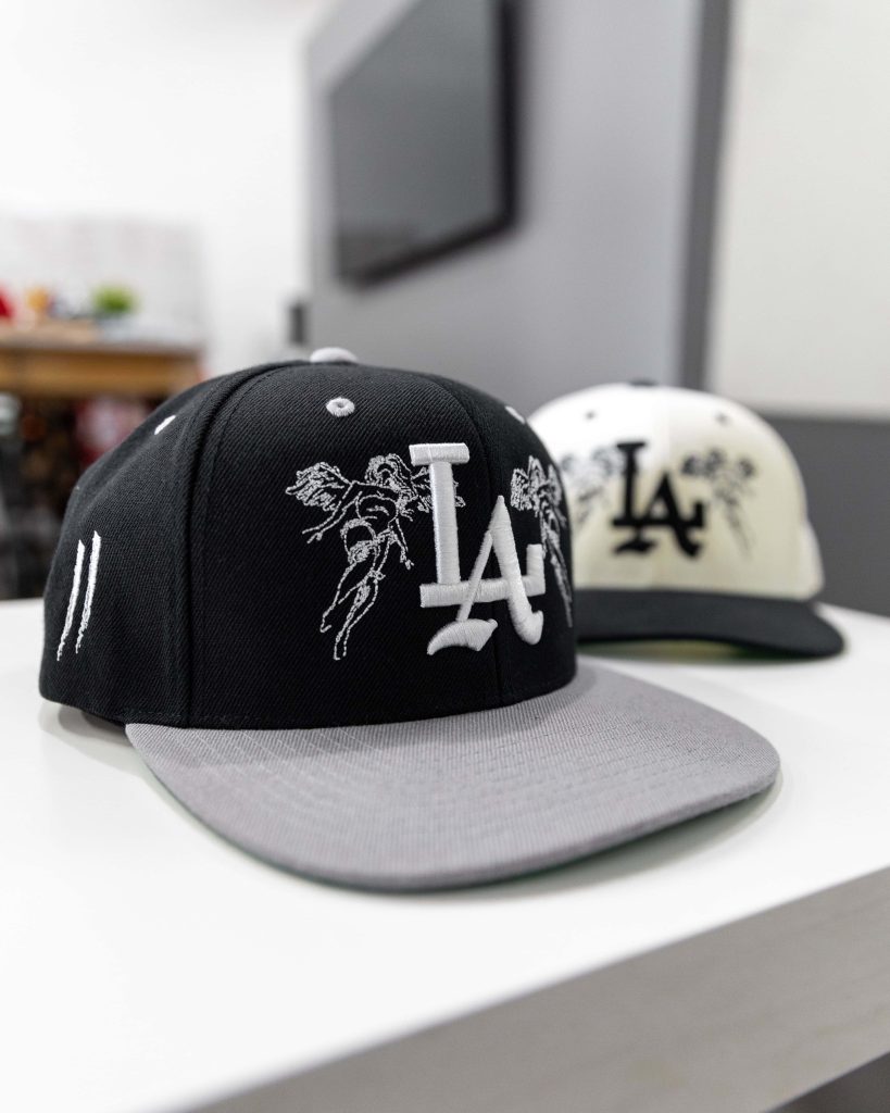 Two custom LA baseball caps with black angel embroidery and cream background urban style Wallpaper