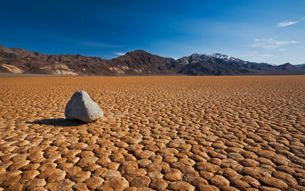 Arid desert landscape with cracked dry lakebed solitary stone and rugged mountains under blue sky Wallpaper
