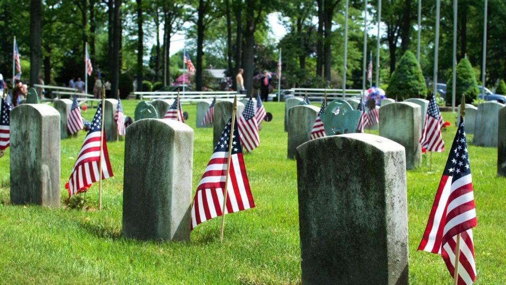 Rows of weathered veterans cemetery headstones with American flags on green grass background Wallpaper