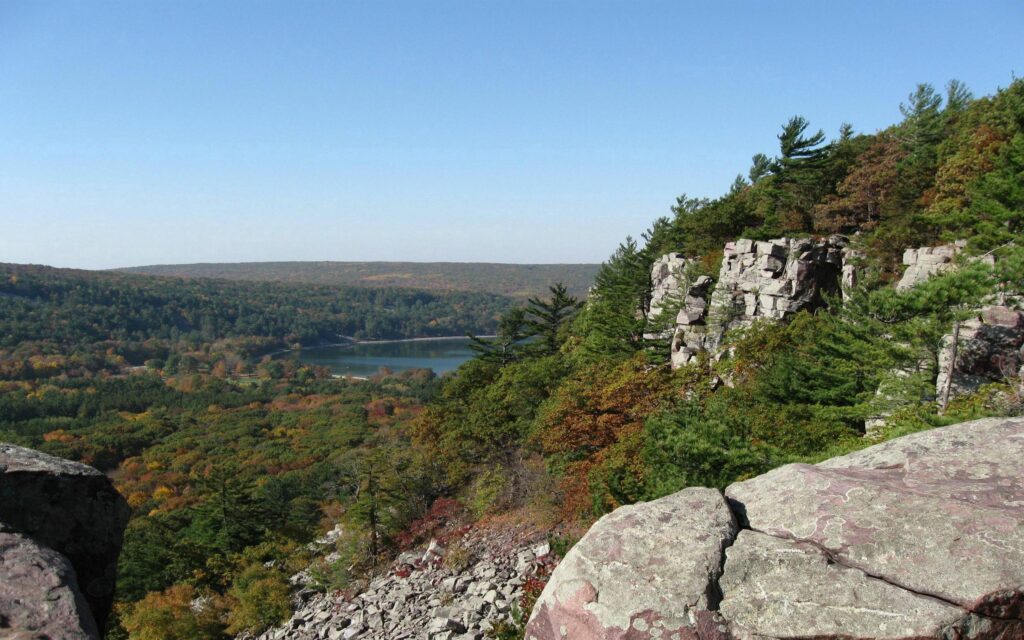 Aerial view of rugged cliffs lush green forests rolling hills and distant lake under clear sky wallpaper