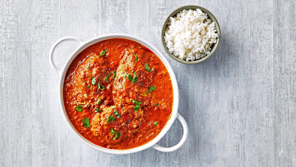 Butter chicken curry in white bowl topped with cilantro beside fluffy white rice on gray background Wallpaper