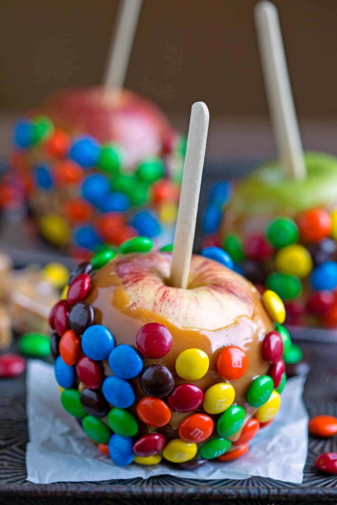 Glossy caramel apples covered in colorful candy pieces on dark background festive wallpaper