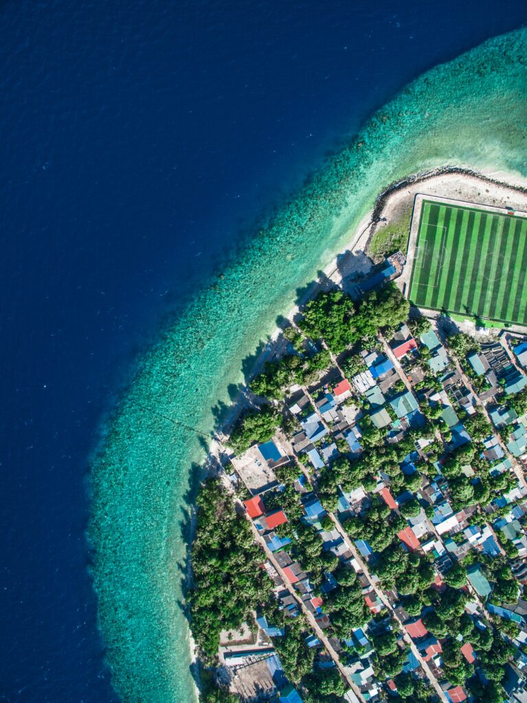 Aerial view of coastal island with colorful rooftops green sports field turquoise reefs and ocean Wallpaper