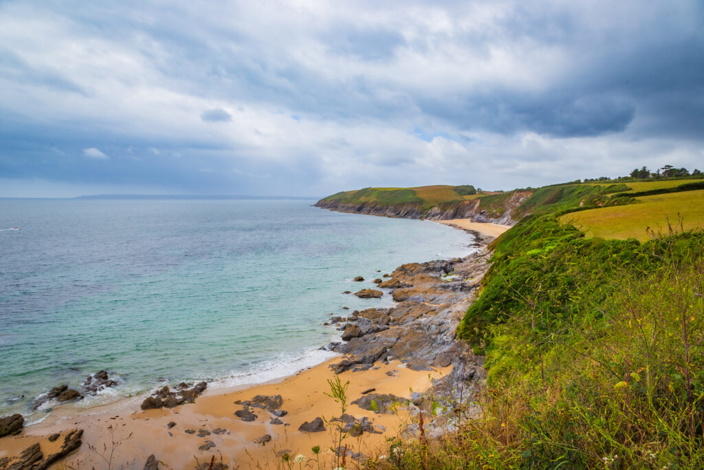 Vibrant coastal wallpaper with rugged stones golden sand blue sea green hills under cloudy sky