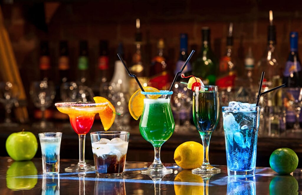 Vibrant bar scene with colorful fruit garnished cocktails in diverse glassware against blurred bottles Wallpaper