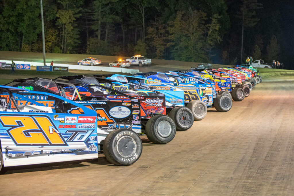 Vibrant dirt track race cars with colorful liveries racing under evening lights and trees backdrop Wallpaper