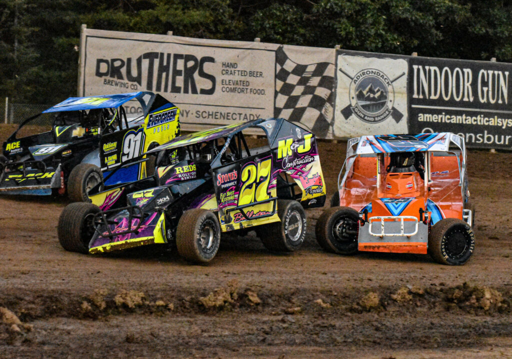 Vibrant dirt track race with sponsor cars speeding on rough earth against green trees wallpaper