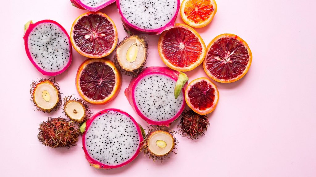 Vibrant flat lay of sliced white dragon fruit blood oranges and rambutans on pastel pink background Wallpaper
