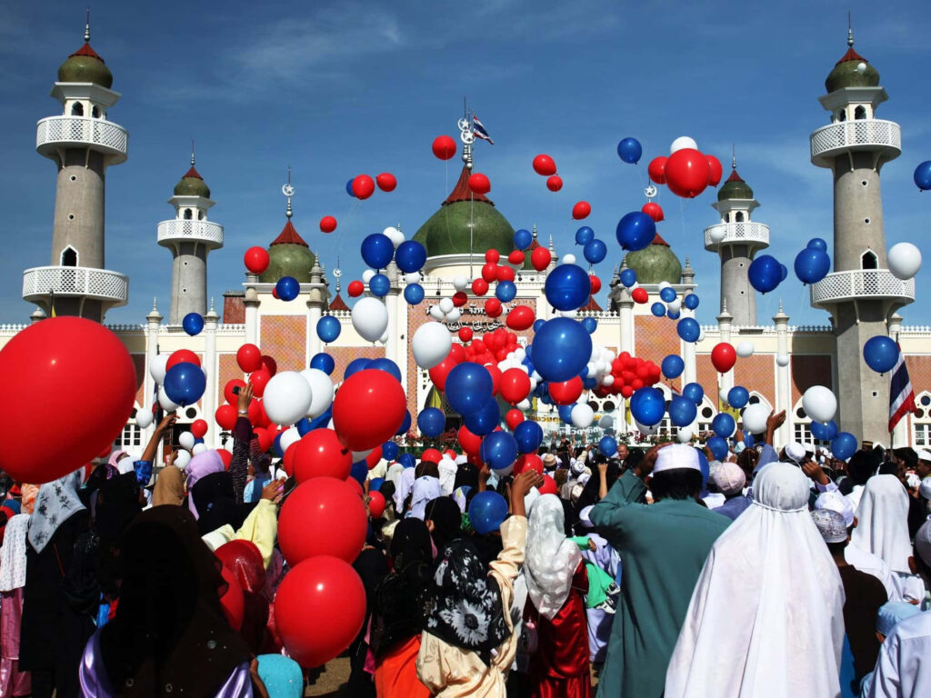 Vibrant Eid celebration crowd at grand mosque with green domes and colorful balloons in sky Wallpaper