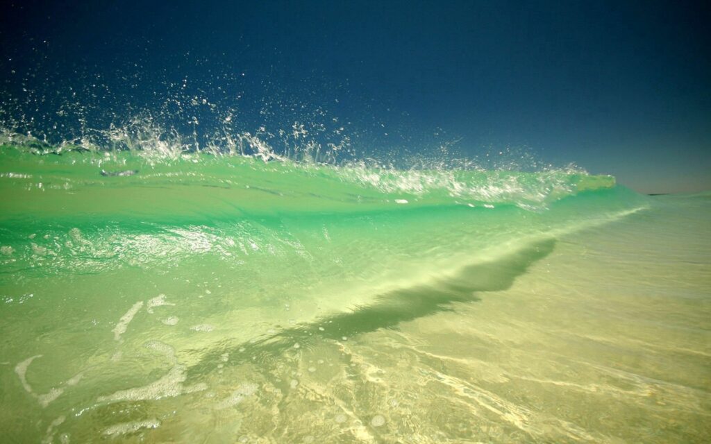 Vivid green ocean wave with sparkling droplets rolling toward coast under deep blue sky wallpaper