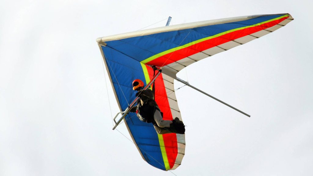 Hang glider with blue red yellow wing banking mid-flight over bright cloudy sky wallpaper background